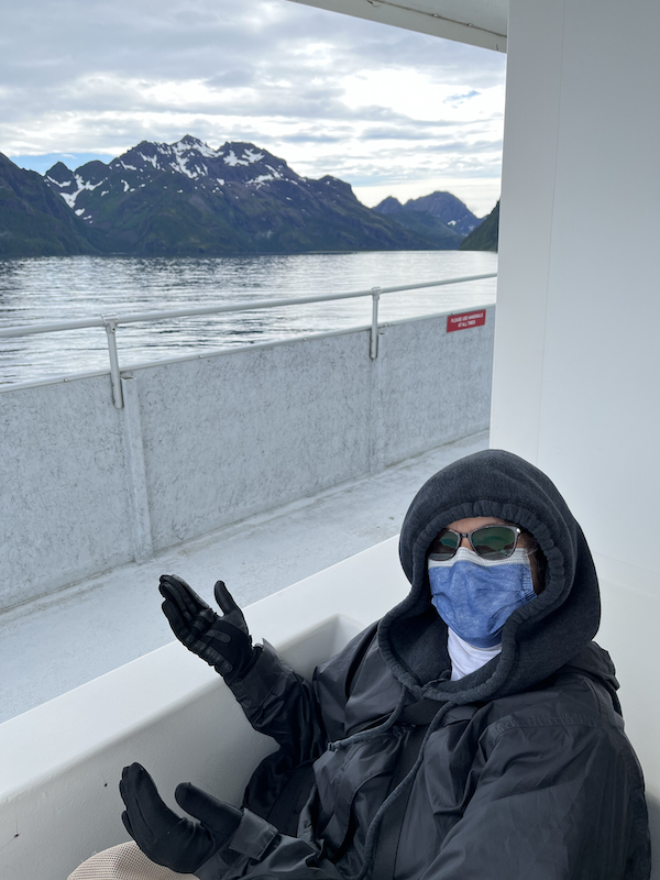 Woman dressed in a hoodie, coat, gloves, sunglasses, and mask on a boat in front of mountains