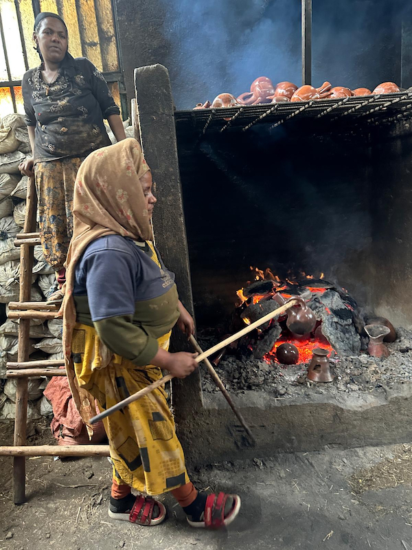 Ethiopian woman removing a clay pot from a fire with a stick through the pot's handle. 